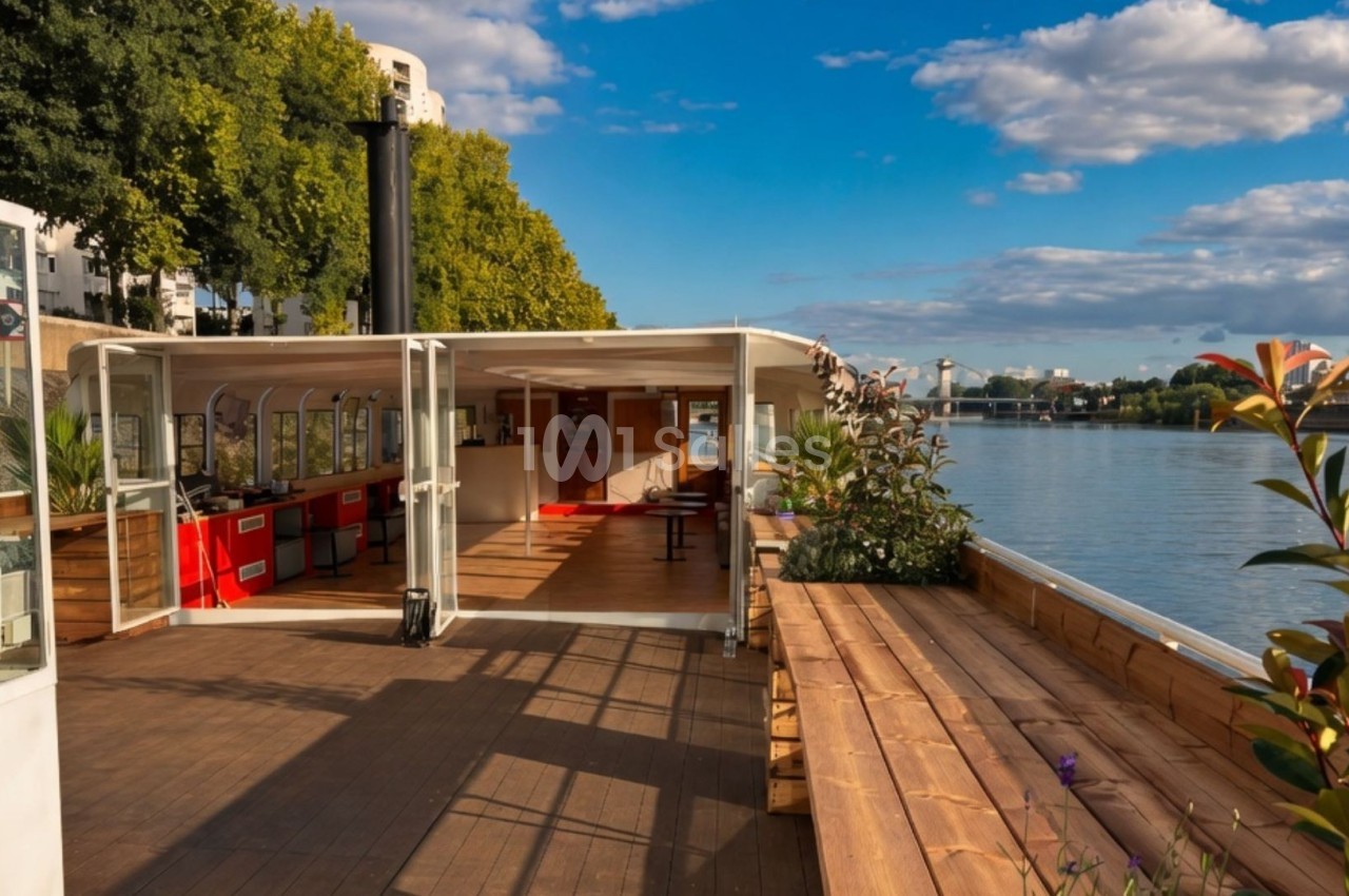Terrasse en bois d'une péniche aménagée avec vue sur une rivière, entourée de végétation et sous un ciel dégagé.