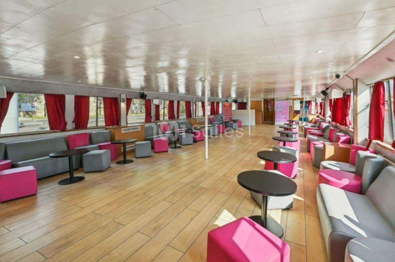 Intérieur d'une salle spacieuse avec parquet, canapés colorés, tables rondes noires et rideaux rouges.