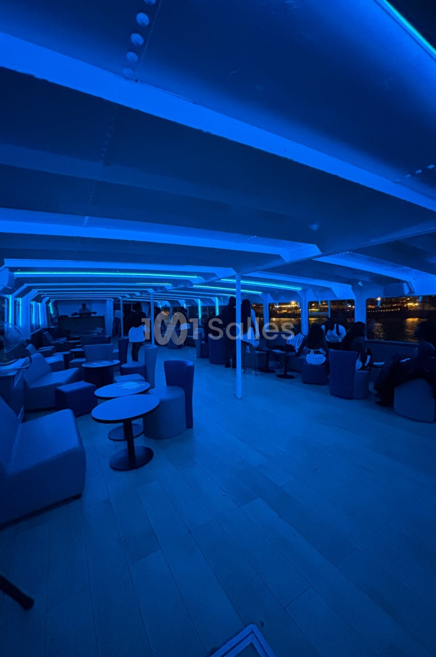 Intérieur d'un bateau avec éclairage bleu, des passagers assis près de tables et des fenêtres donnant sur l'eau.