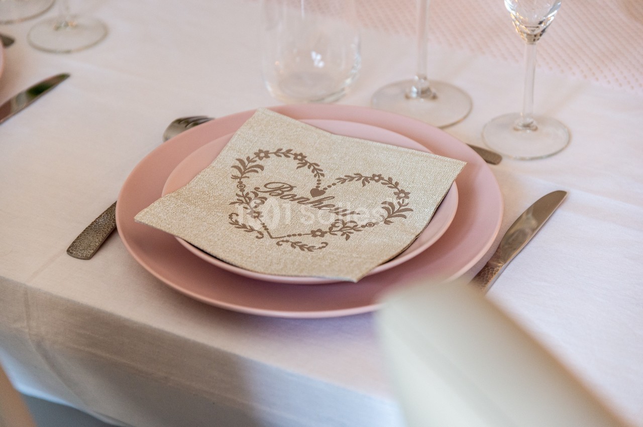 Serviette décorative avec le mot ’Bonheur’ posée sur une assiette rose, entourée de couverts et de verres.
