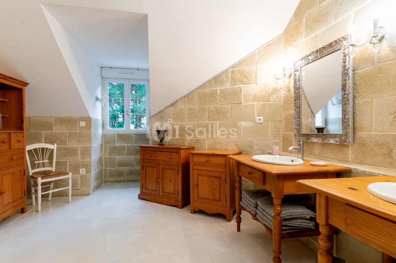 Salle de bain lumineuse avec meubles en bois, miroir encadré, murs en pierre beige et fenêtre donnant sur des arbres.