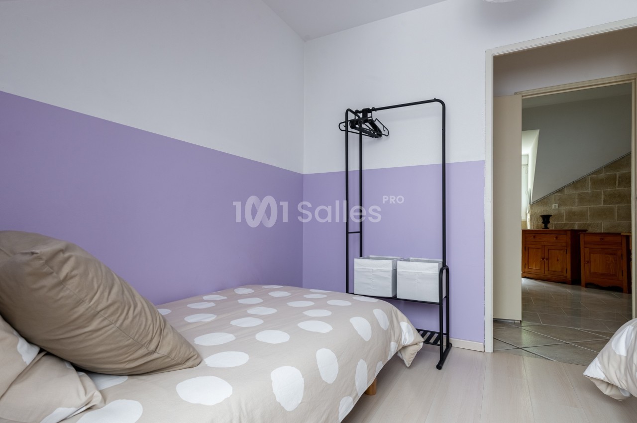 Chambre avec deux lits simples, murs violets, portant noir avec cintres et vue sur une pièce adjacente.