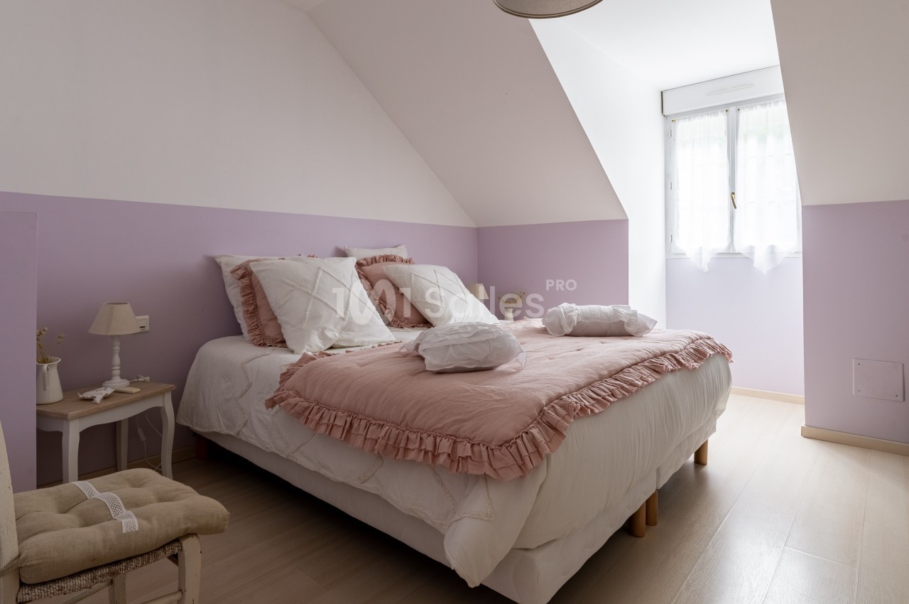 Chambre lumineuse avec lit double, draps roses et blancs, murs lilas, fenêtre avec rideaux légers et mobilier simple.