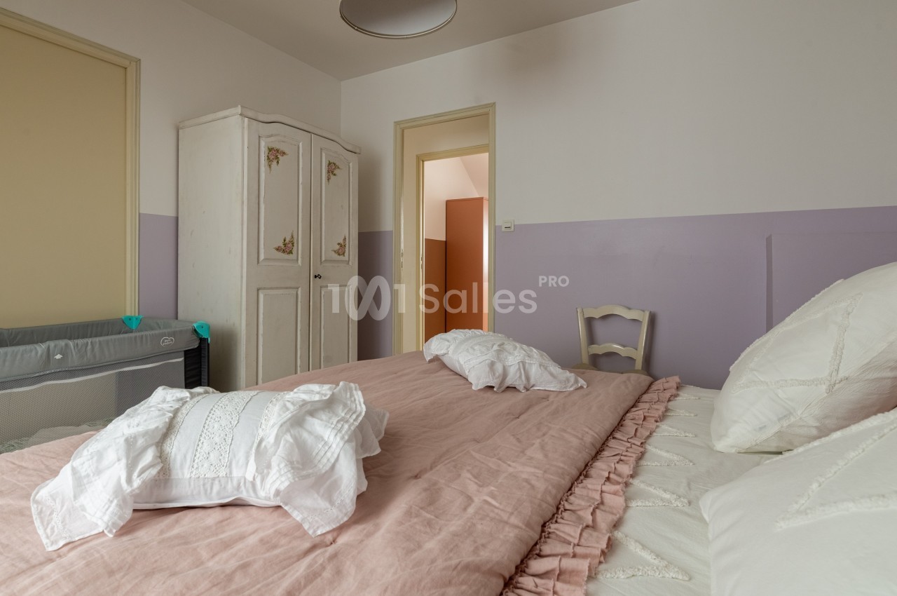 Lit double avec draps roses dans une chambre lumineuse, armoire blanche et lit bébé en arrière-plan.