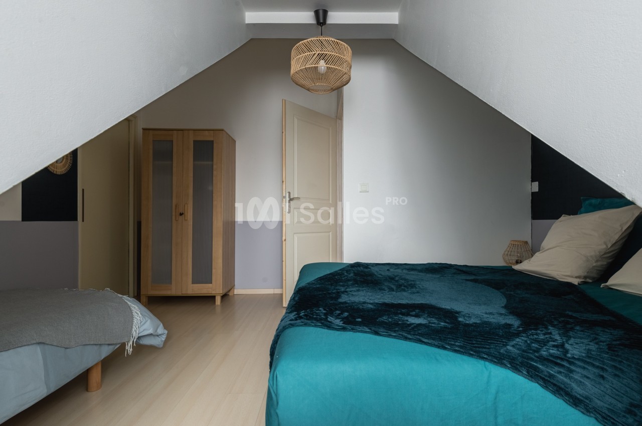 Chambre mansardée avec lit double, lit simple, armoire en bois et suspension en rotin.