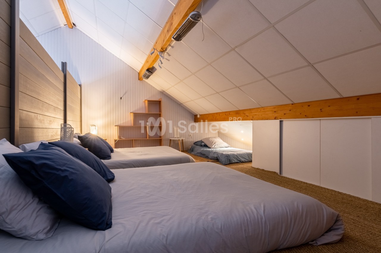 Chambre mansardée avec trois lits, poutres apparentes, éclairage doux et étagères en bois dans un coin.