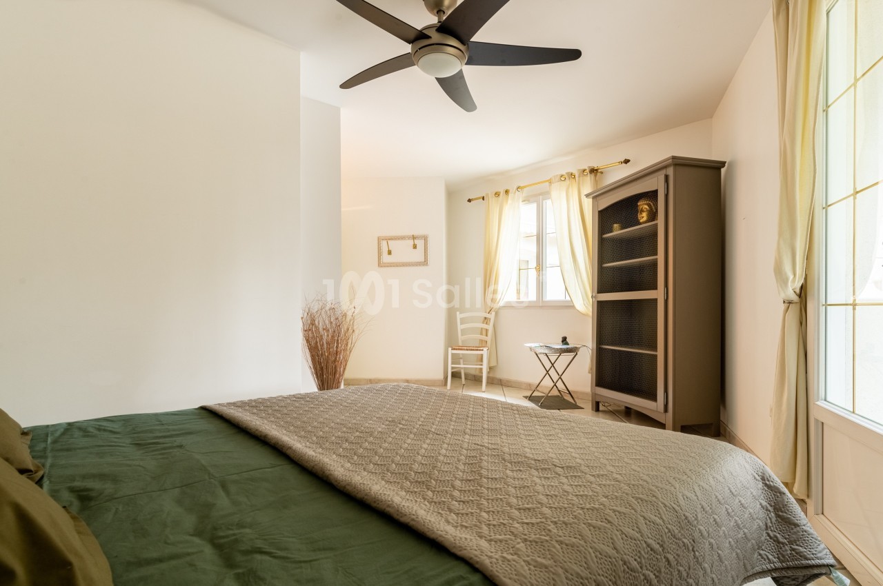 Chambre lumineuse avec lit double, armoire, chaise et table près d'une fenêtre aux rideaux clairs.
