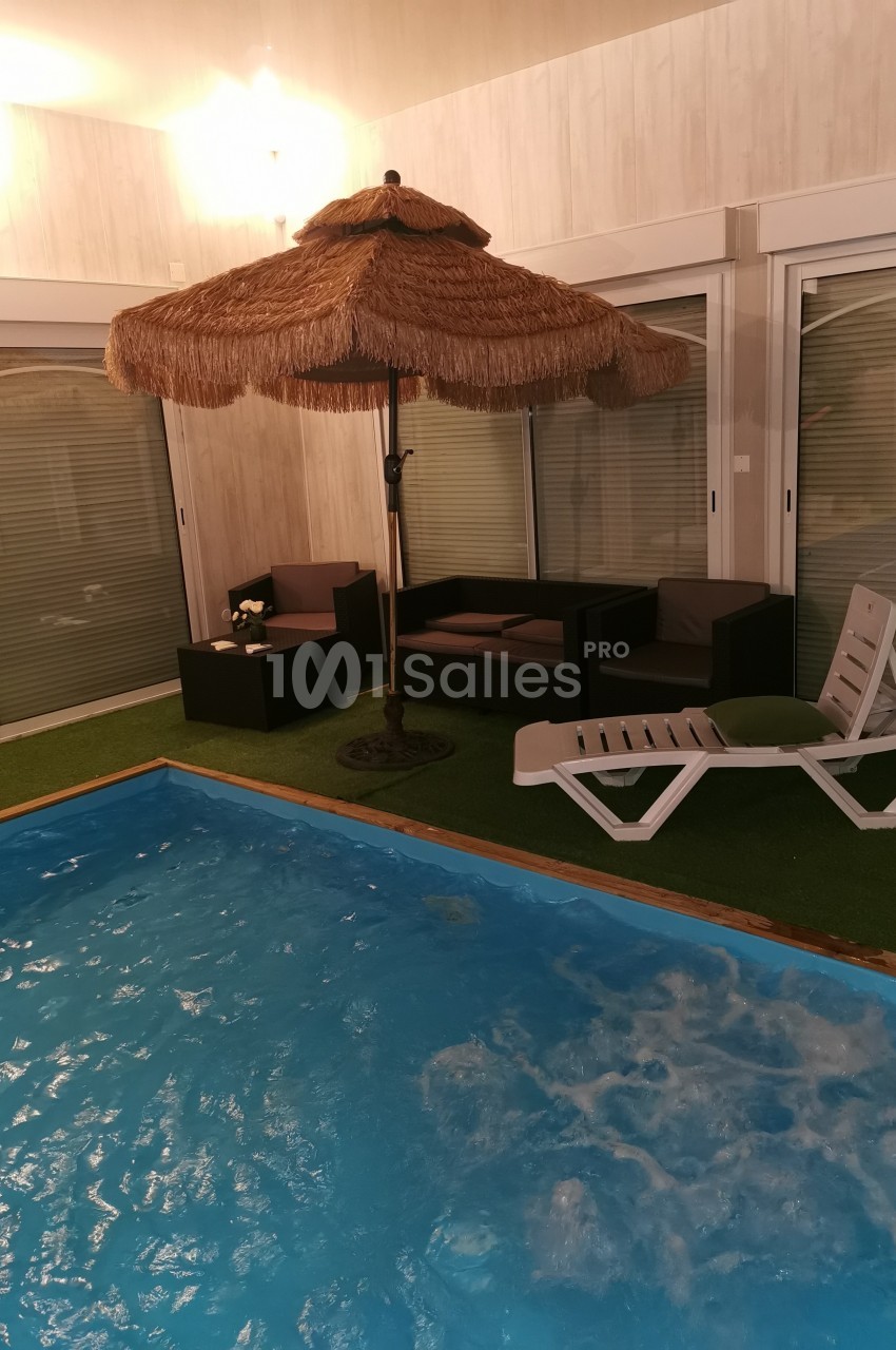 Espace intérieur avec piscine, parasol en paille, transat blanc et mobilier de jardin sur gazon artificiel.