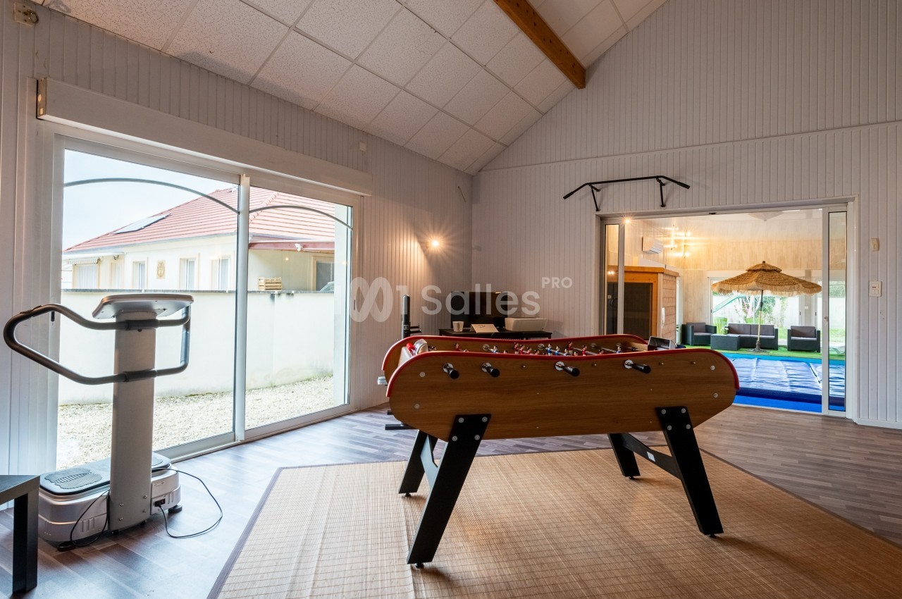Salle de loisirs avec baby-foot, équipement de fitness et vue sur un espace extérieur et une piscine couverte.