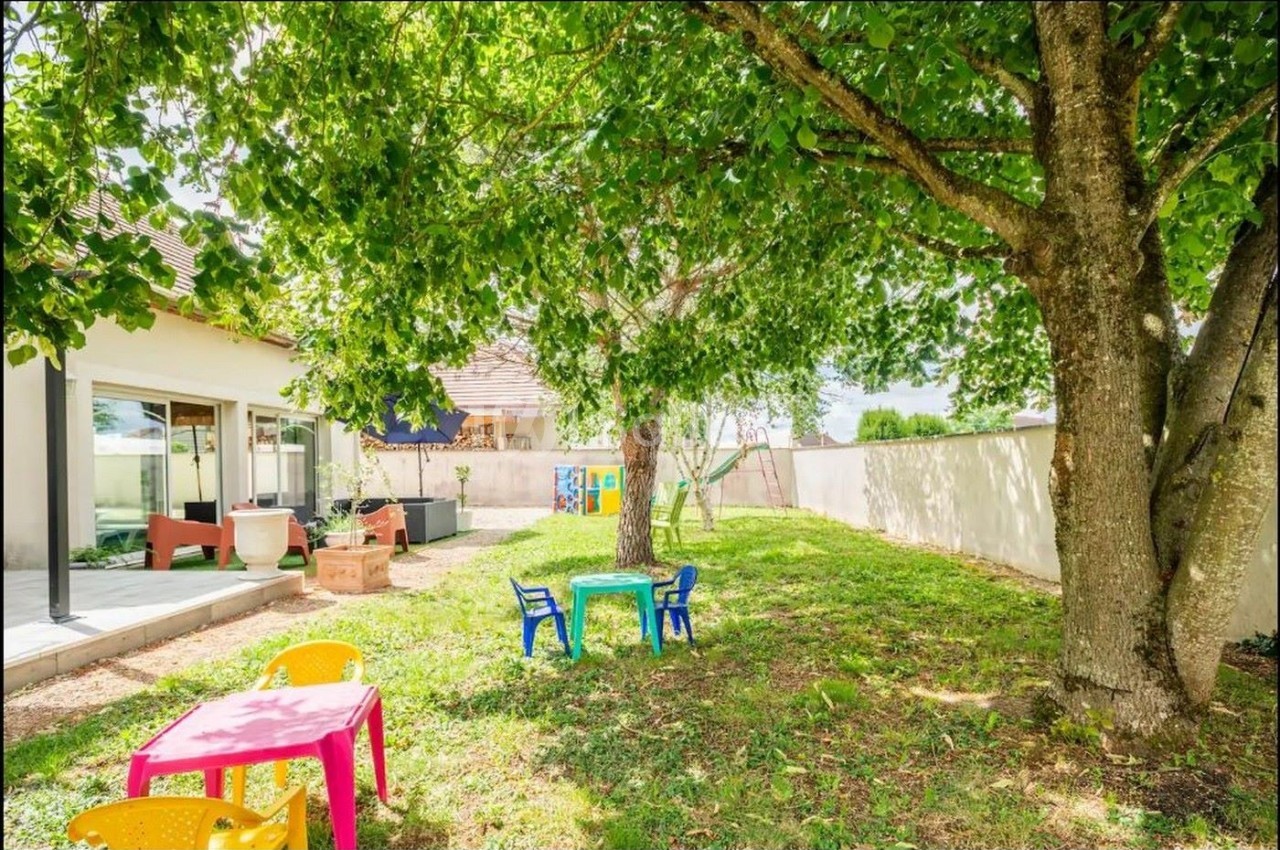 Jardin avec pelouse, arbres, tables et chaises colorées pour enfants, adjacent à une maison avec terrasse.