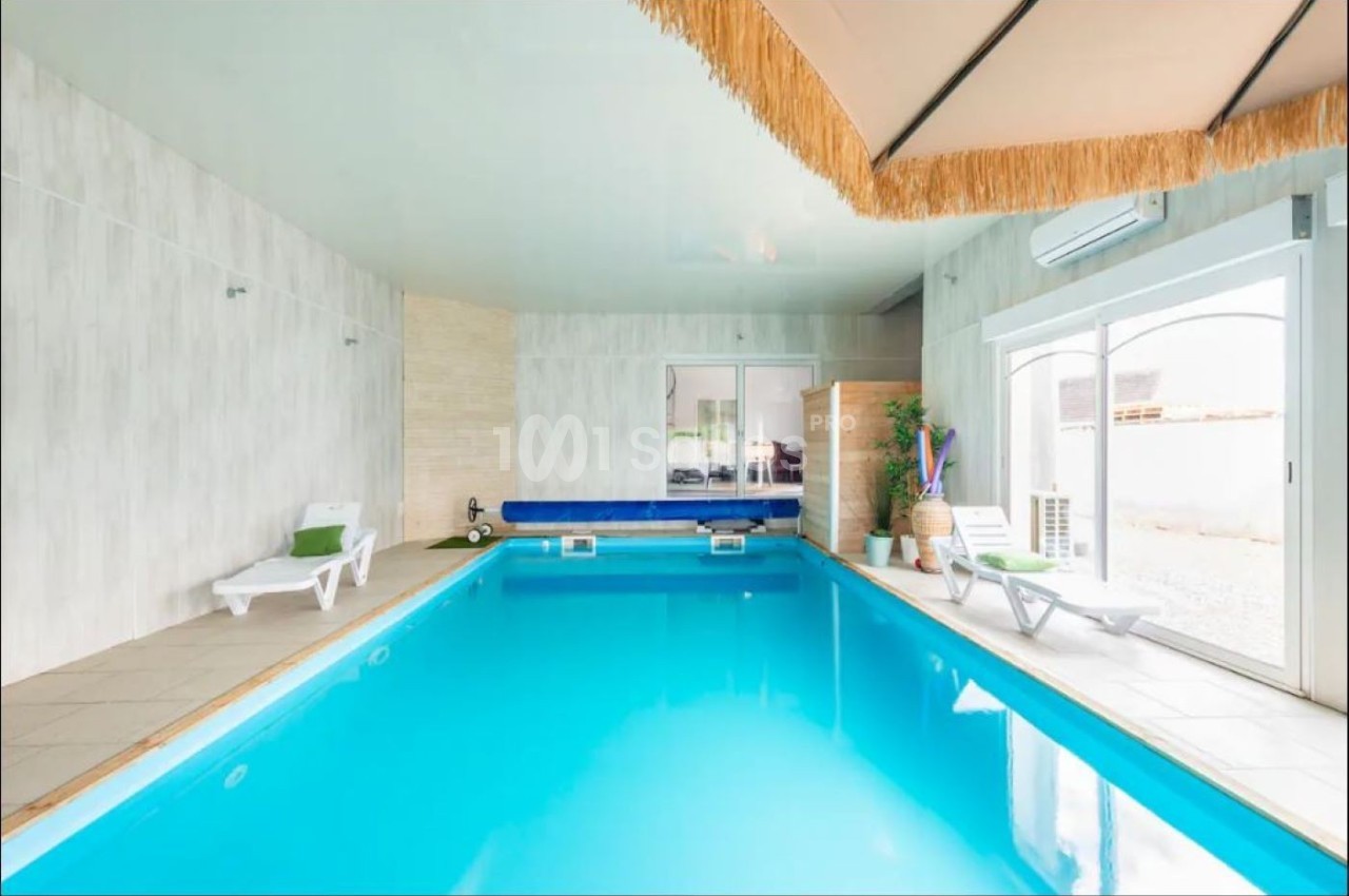 Piscine intérieure avec eau turquoise, entourée de chaises longues blanches et décorée de plantes et d'un parasol.