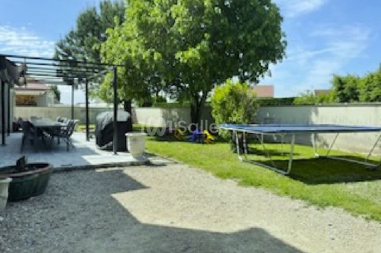 Jardin avec terrasse ombragée, trampoline, pelouse, arbre et clôture en arrière-plan sous un ciel dégagé.