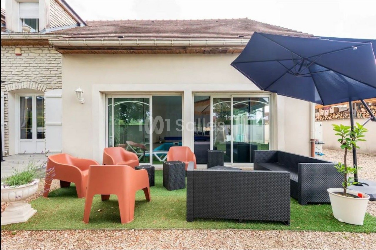 Terrasse avec mobilier de jardin en résine tressée et chaises orange, parasol bleu, devant une maison en pierre.