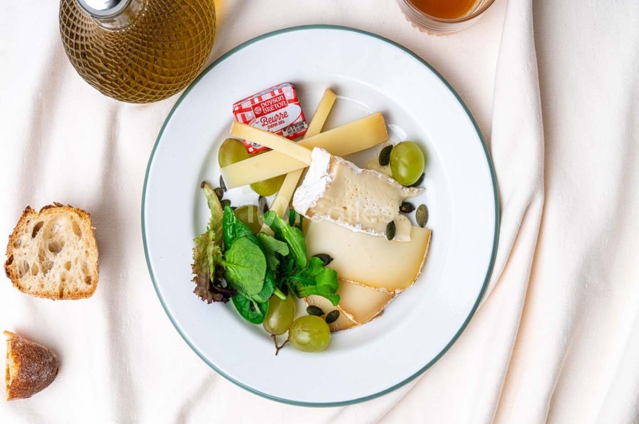 Assiette de fromages variés avec raisins, salade verte et graines, accompagnée de pain et d'huile d'olive.