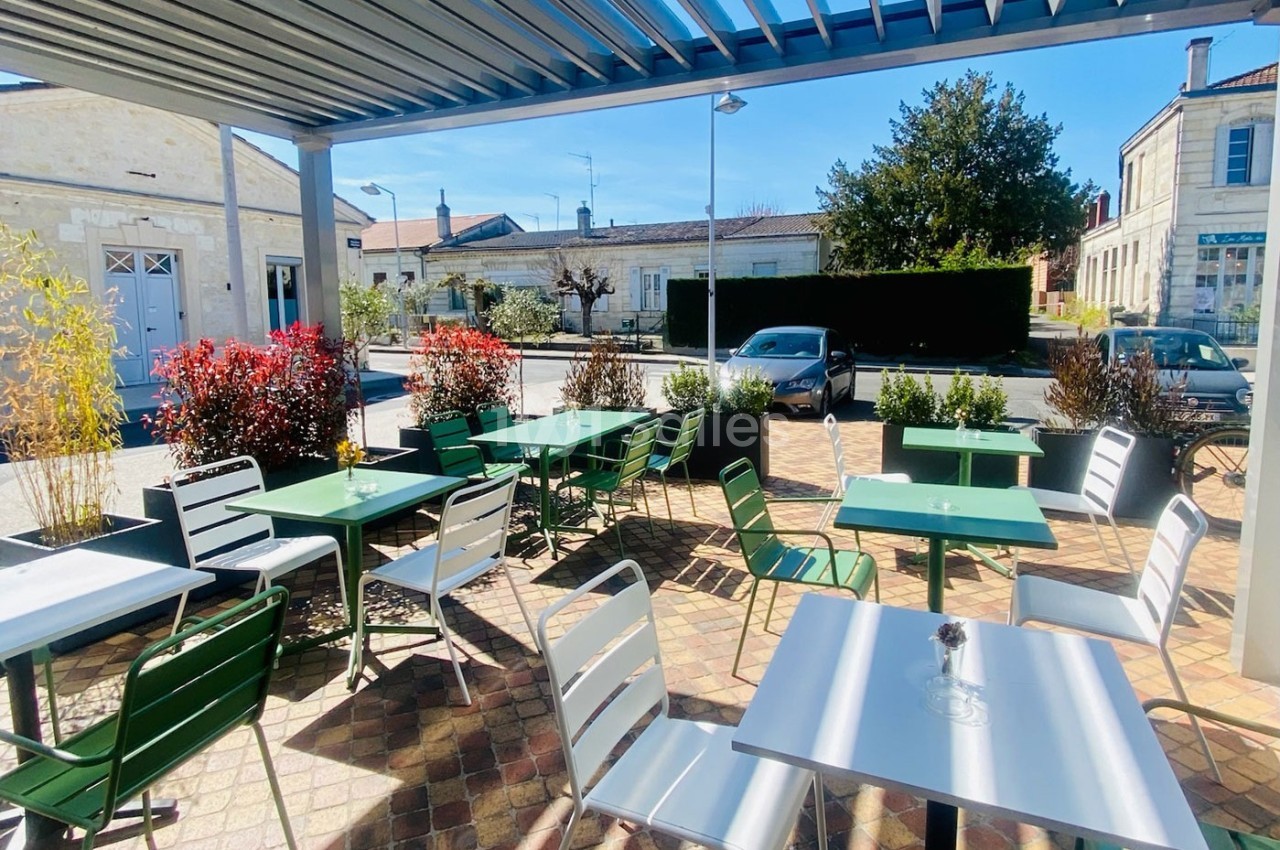 Terrasse extérieure avec tables et chaises colorées sous une pergola, entourée de plantes et donnant sur une rue calme.