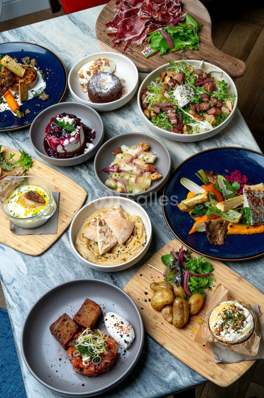 Table garnie de plats variés : légumes grillés, viandes, fromages, houmous, pommes de terre et salades colorées.