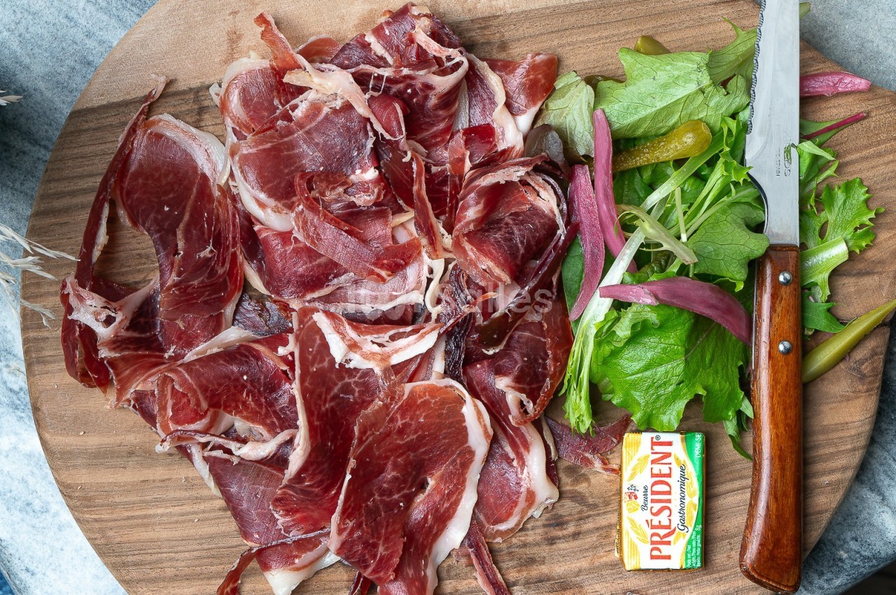Assiette de jambon cru tranché avec salade verte, cornichons, beurre et couteau sur une planche en bois.