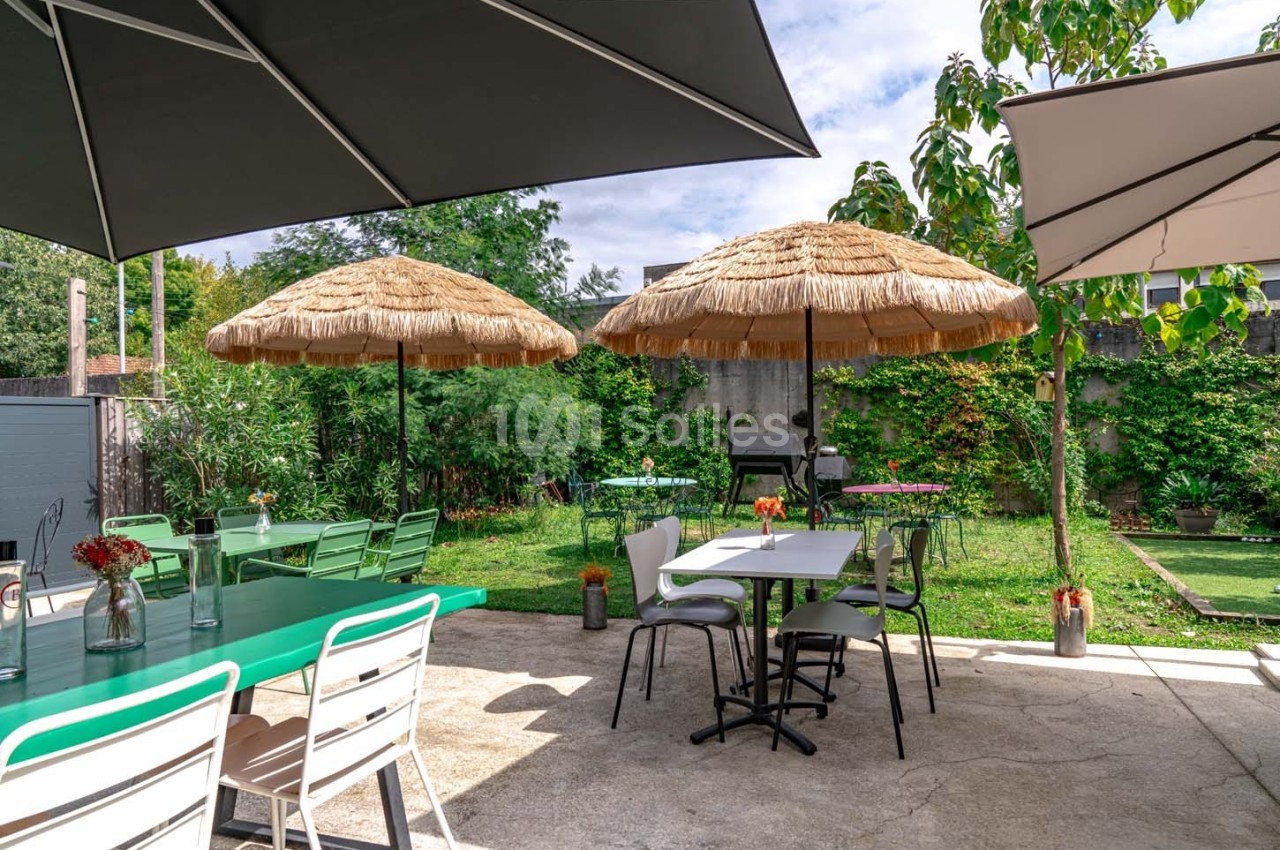 Terrasse extérieure avec tables, chaises, parasols en paille et végétation environnante sous un ciel partiellement nuageux.