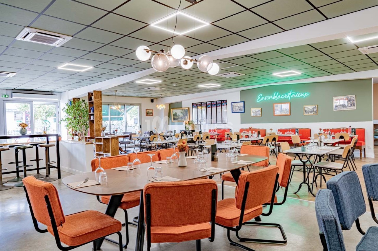Salle de restaurant moderne avec tables dressées, chaises colorées et décoration lumineuse au mur.