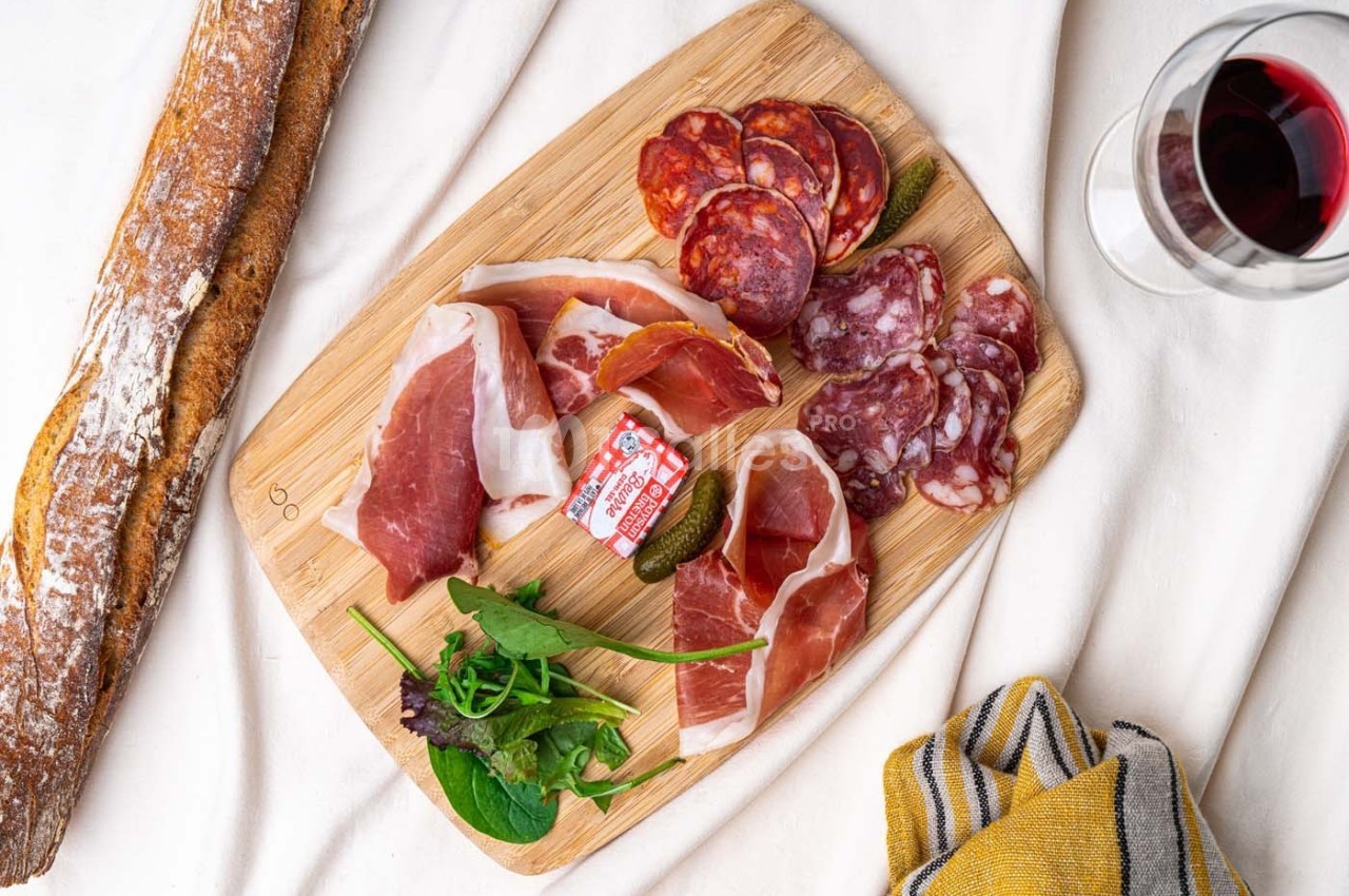 Planche en bois garnie de charcuterie, cornichons, salade, fromage, avec une baguette et un verre de vin rouge à côté.