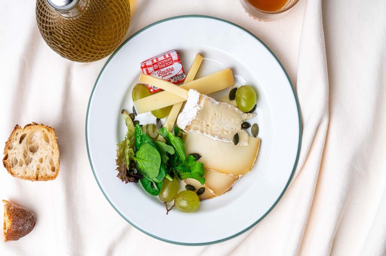 Assiette de fromages variés avec raisins, salade verte et graines, accompagnée de pain et d'huile d'olive.