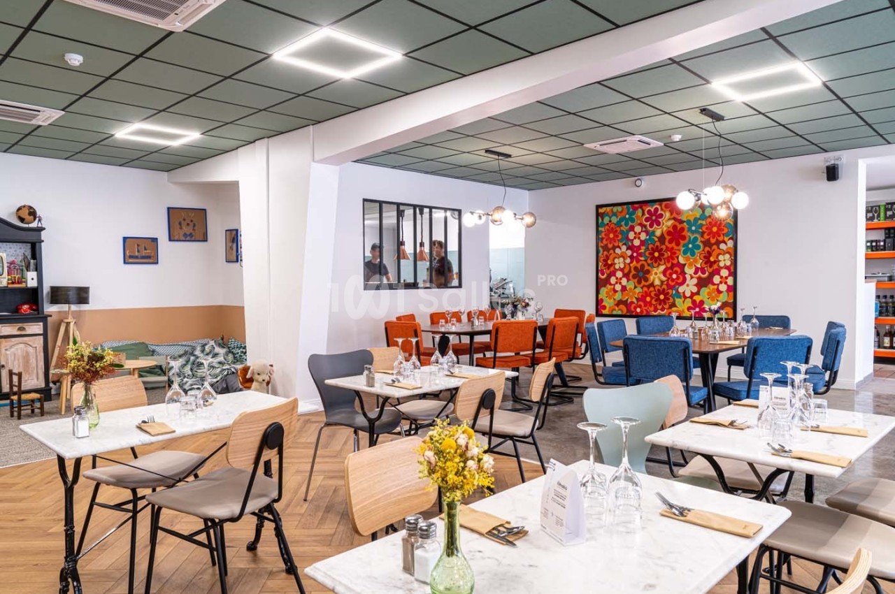 Salle de restaurant lumineuse avec tables dressées, chaises variées et décoration colorée sur les murs.