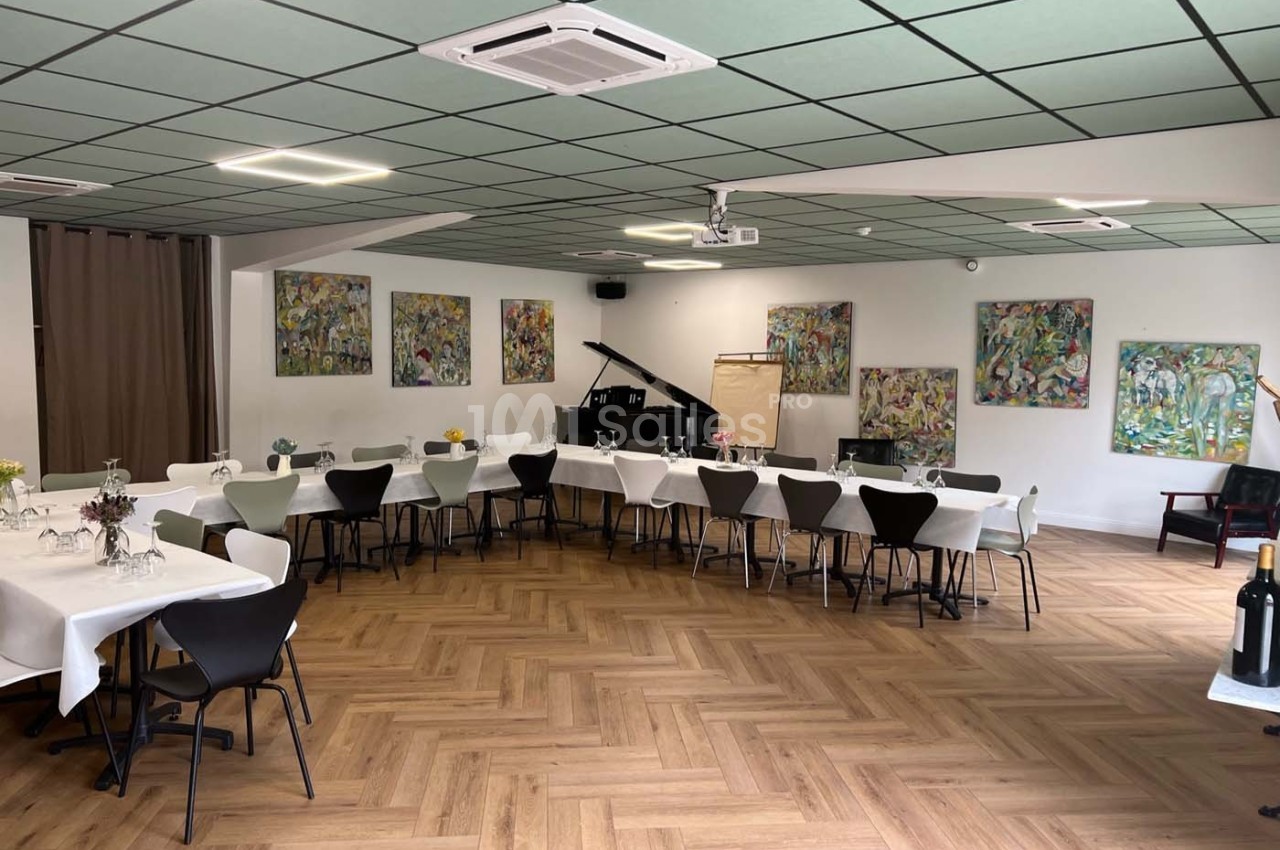 Salle lumineuse avec parquet, tables dressées, chaises noires et blanches, piano à queue et tableaux colorés aux murs.