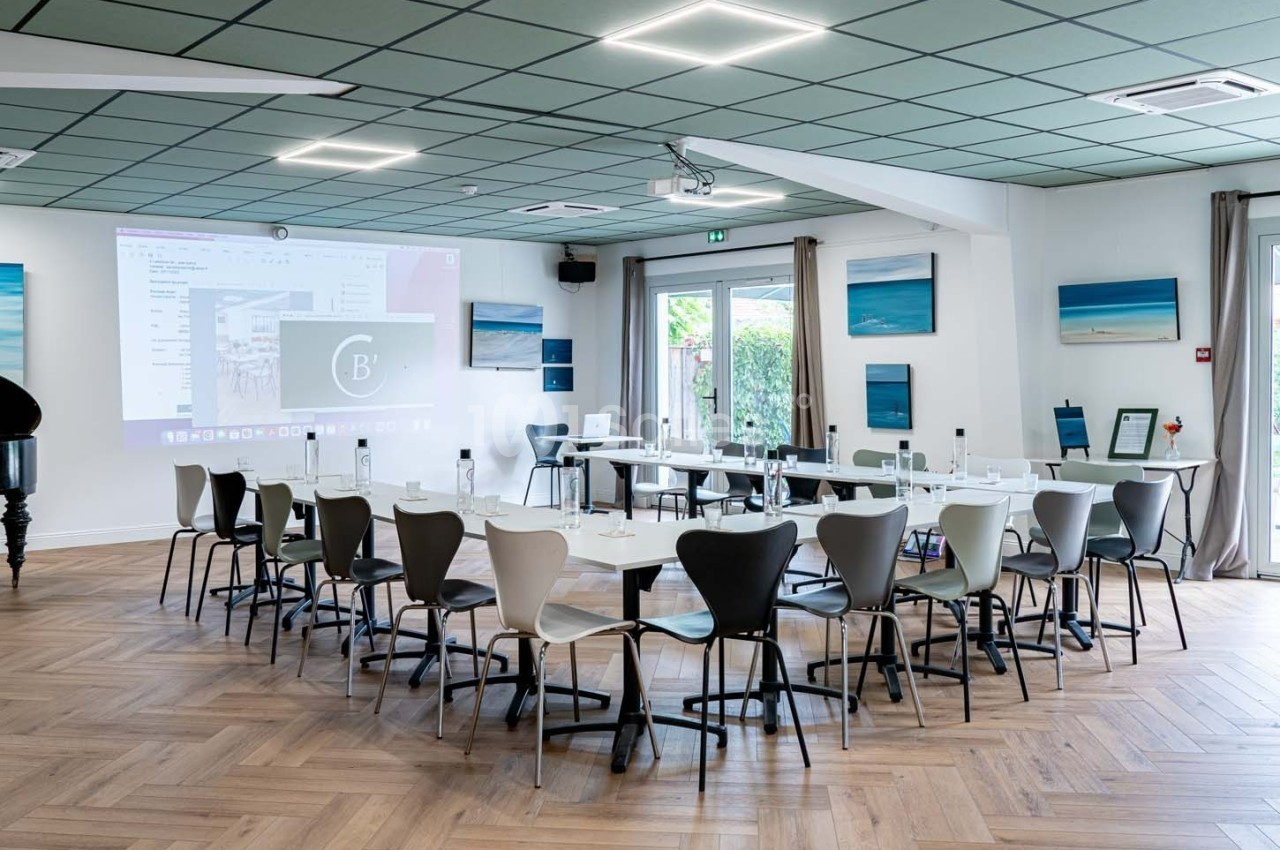 Salle de réunion lumineuse avec tables disposées en U, chaises modernes, écran de projection et décorations murales.