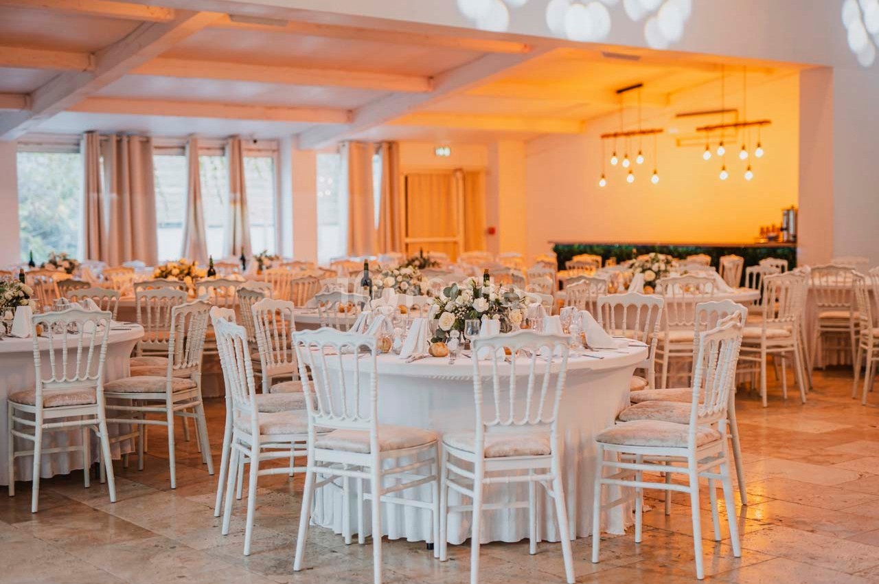 Salle de réception décorée avec des tables rondes, nappes blanches, chaises élégantes et éclairage chaleureux.