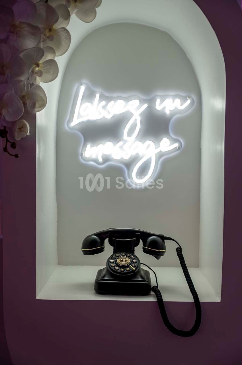 Téléphone rétro noir posé sous un néon blanc indiquant ’Laissez un message’, entouré d'orchidées blanches.