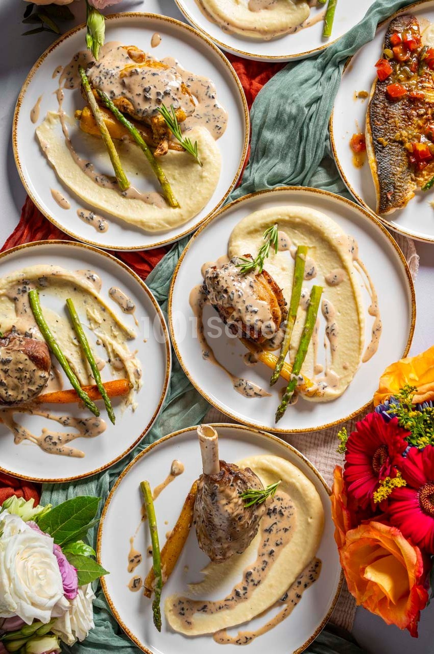 Assiettes garnies de purée, asperges, viande ou poisson, nappées de sauce, disposées sur une table décorée de fleurs.