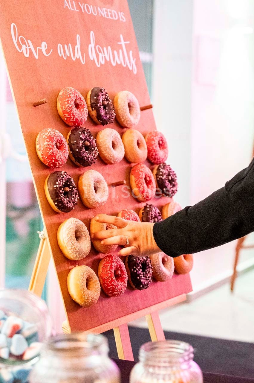 Mur en bois avec des donuts accrochés, une main en saisit un, pots de bonbons visibles au premier plan.