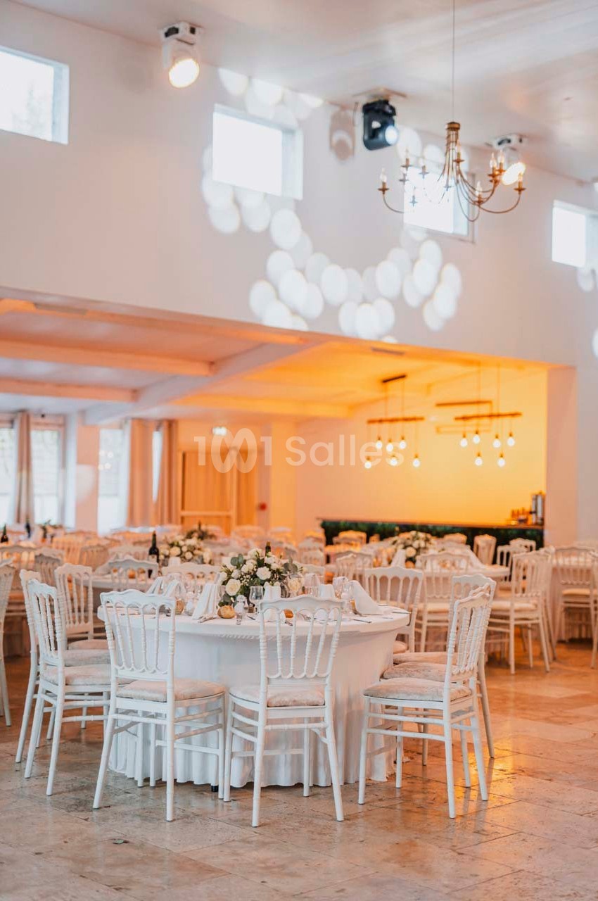 Salle de réception lumineuse avec tables rondes décorées de fleurs blanches et chaises blanches élégantes.