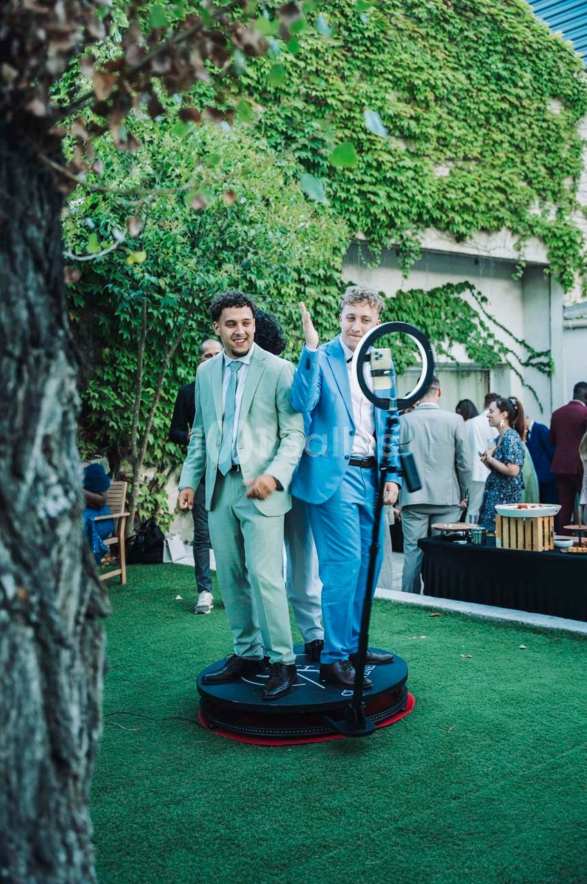 Deux hommes en costume posent sur une plateforme tournante dans un jardin, entourés de verdure et de convives.