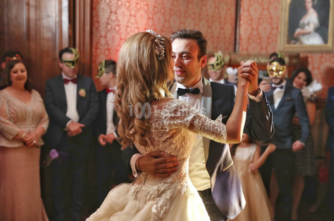 Un couple danse lors d'une réception élégante, entouré d'invités en tenue de soirée et portant des masques.