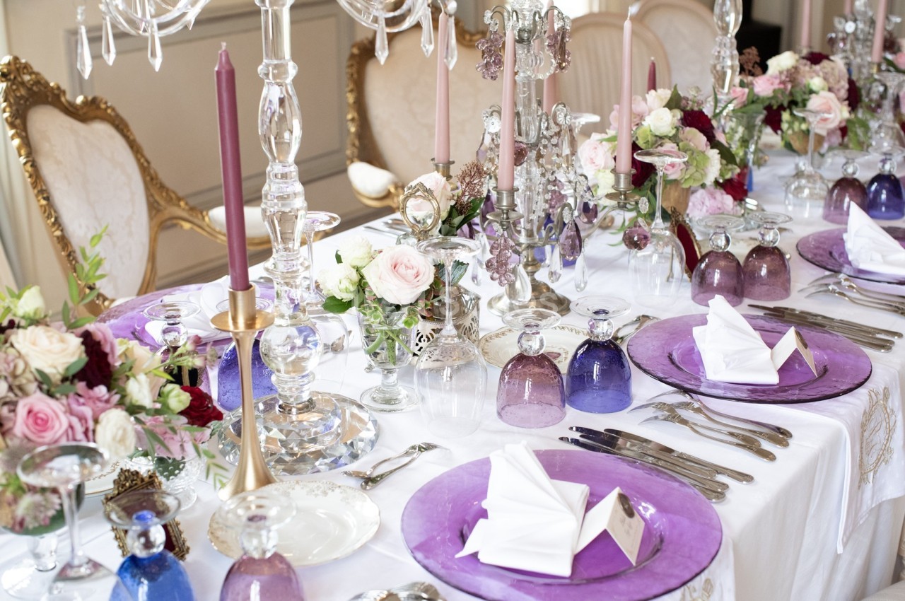 Table élégamment dressée avec assiettes violettes, verres colorés, chandeliers et arrangements floraux.