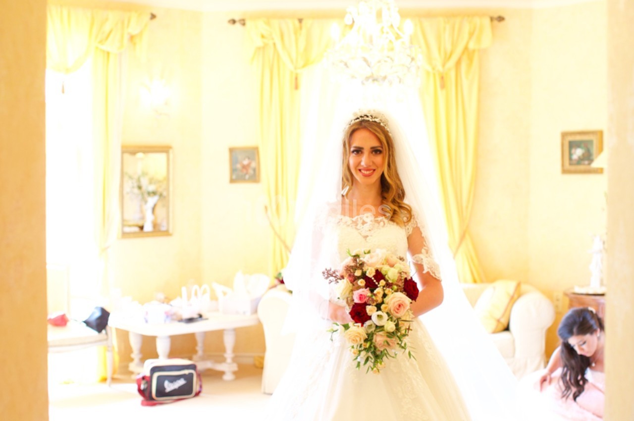 Une mariée souriante en robe blanche tient un bouquet de fleurs dans une pièce lumineuse aux tons jaunes.