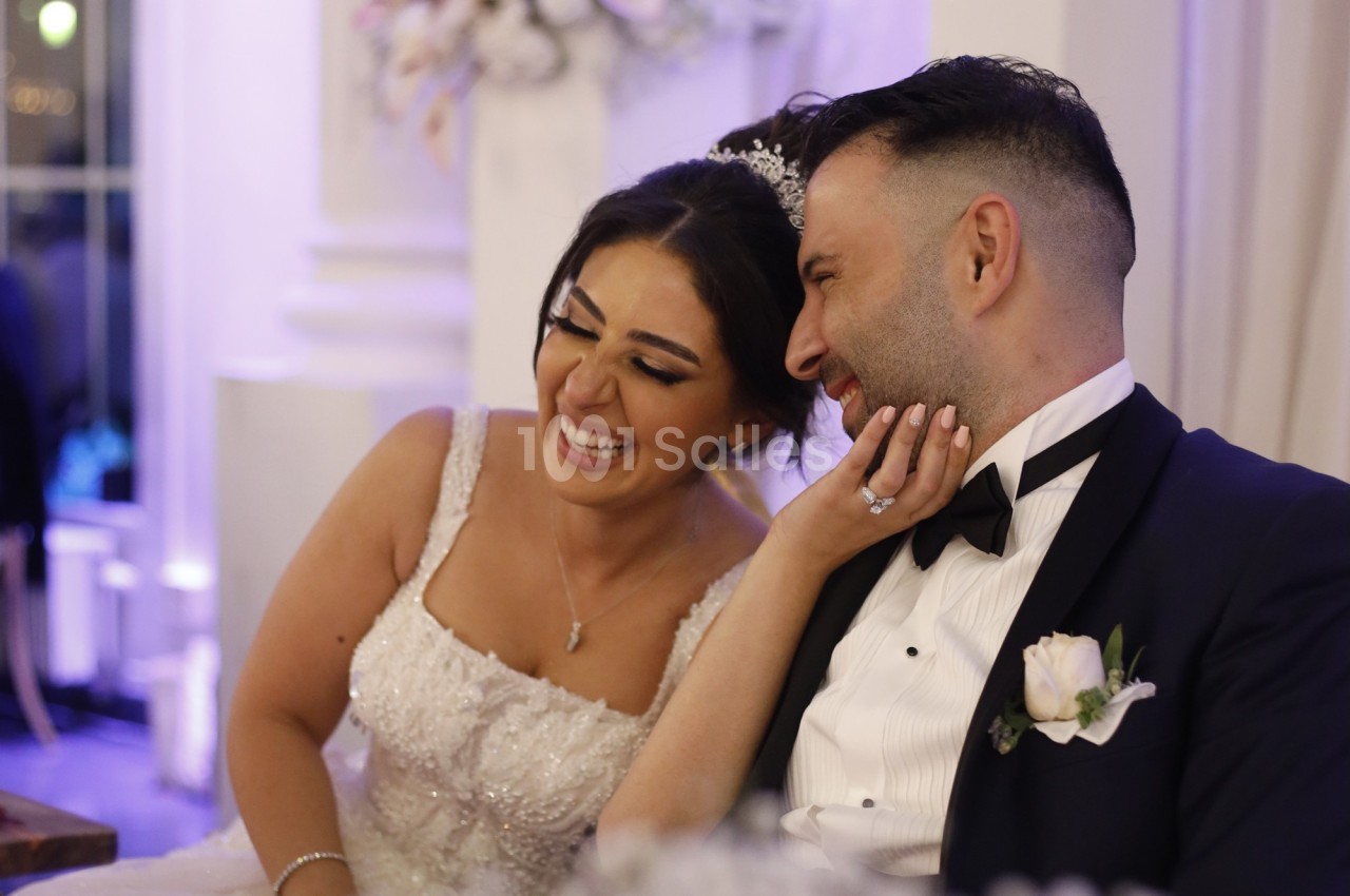 Un couple en tenue de mariage sourit et partage un moment complice dans un décor intérieur élégant.
