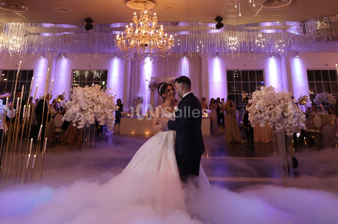 Un couple danse leur première danse de mariage dans une salle élégante éclairée de lumières violettes et dorées.