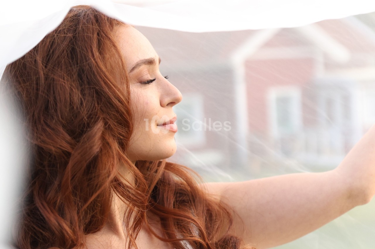 Femme aux cheveux roux souriant légèrement, vue de profil, sous un voile blanc avec des maisons floues en arrière-plan.