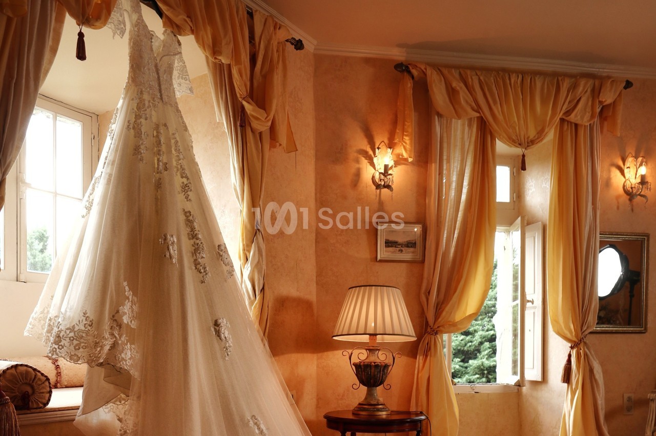 Robe de mariée en dentelle suspendue près d'une fenêtre dans une pièce lumineuse aux rideaux dorés.