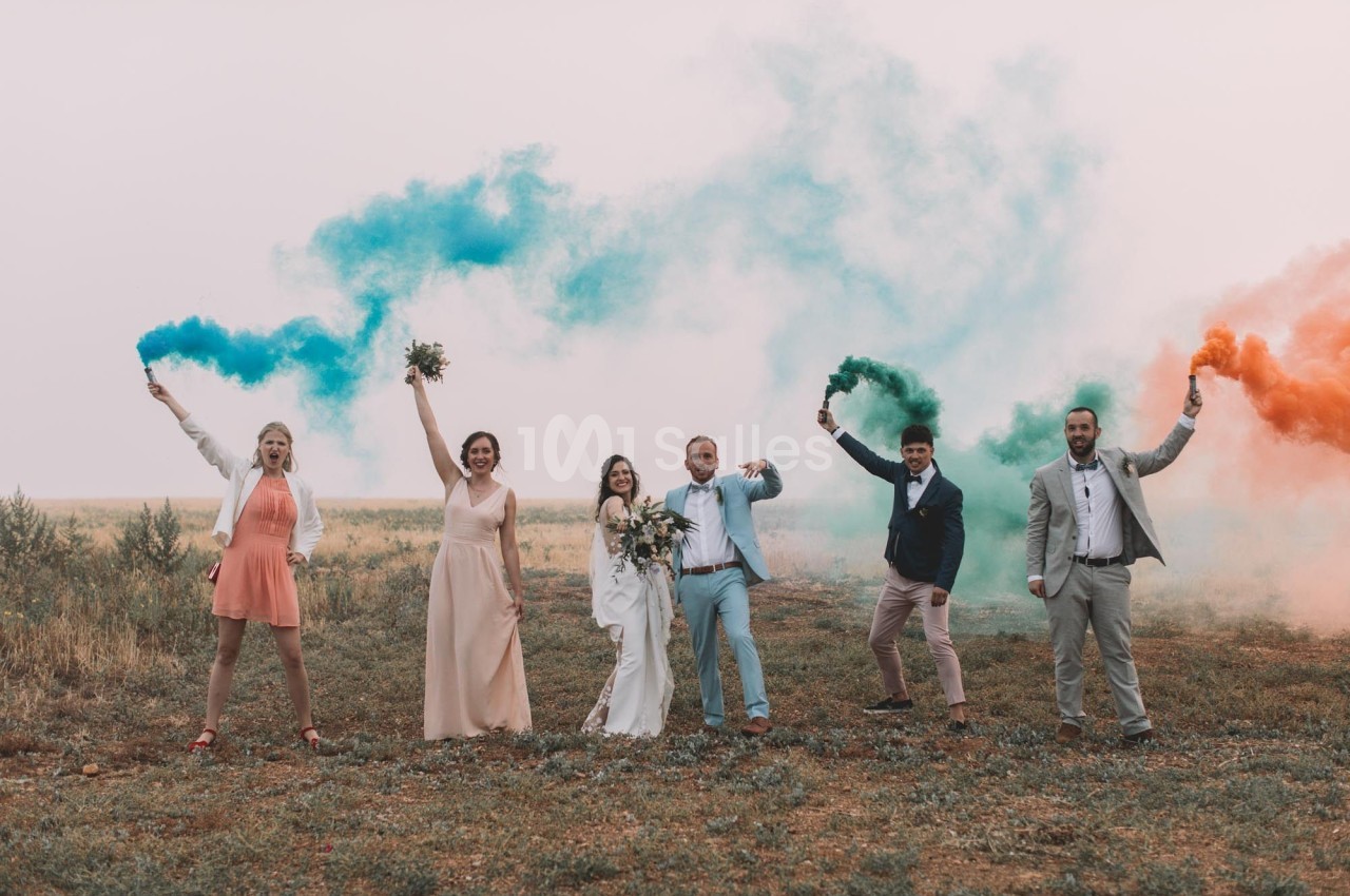 Un groupe de personnes en tenue de cérémonie tient des fumigènes colorés dans un champ ouvert.