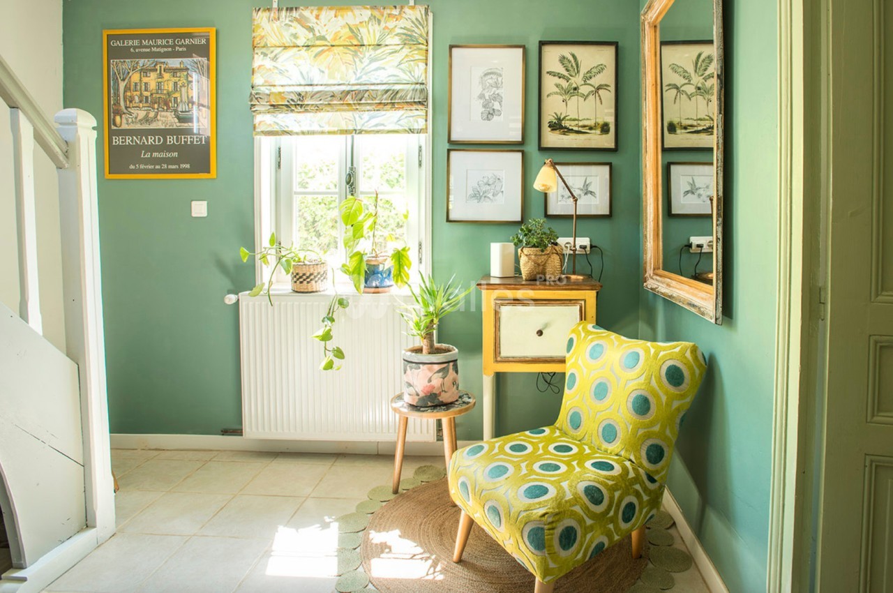 Coin de salon lumineux avec fauteuil jaune à motifs, plantes en pot, mur vert décoré de cadres et fenêtre avec store imprimé.