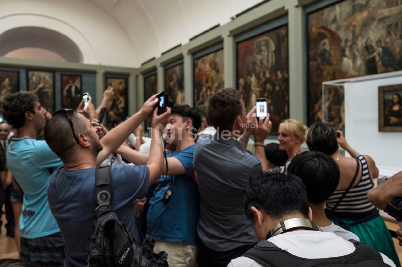 Des visiteurs photographient une œuvre d'art dans une galerie bondée, entourés de peintures exposées sur les murs.