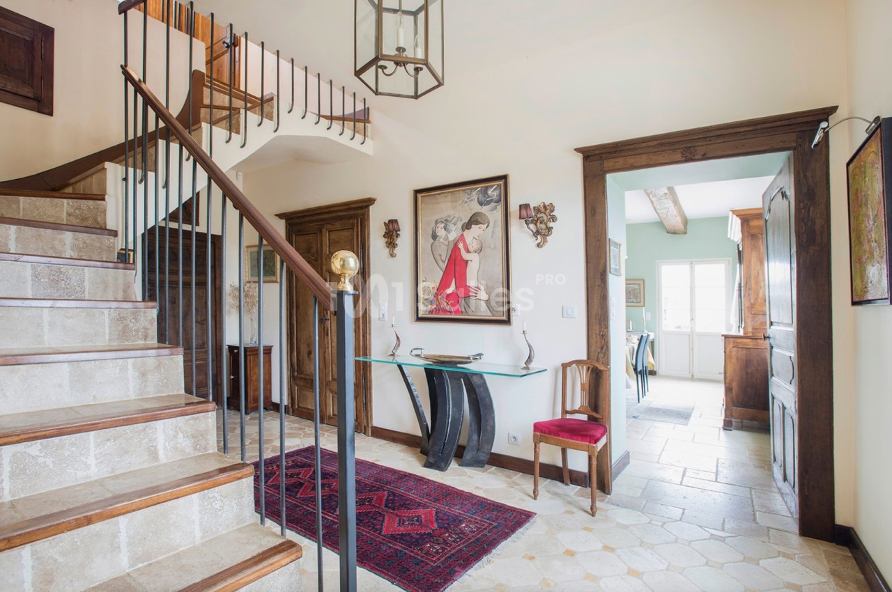 Entrée lumineuse avec escalier en pierre, tapis rouge, meubles en bois et accès à une cuisine au fond.