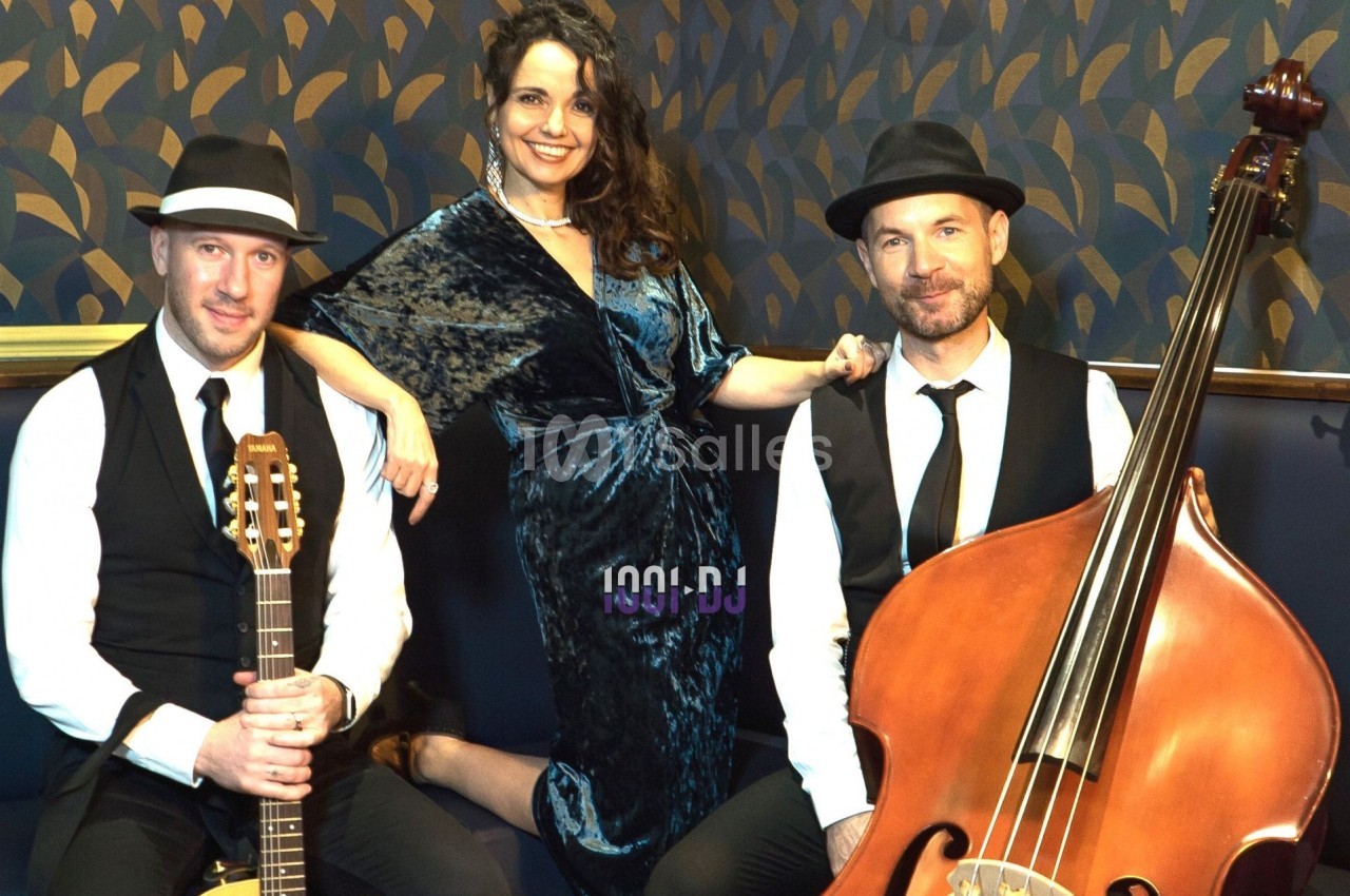 Un trio musical pose avec une guitare, une contrebasse et une femme en robe bleue dans un décor élégant.