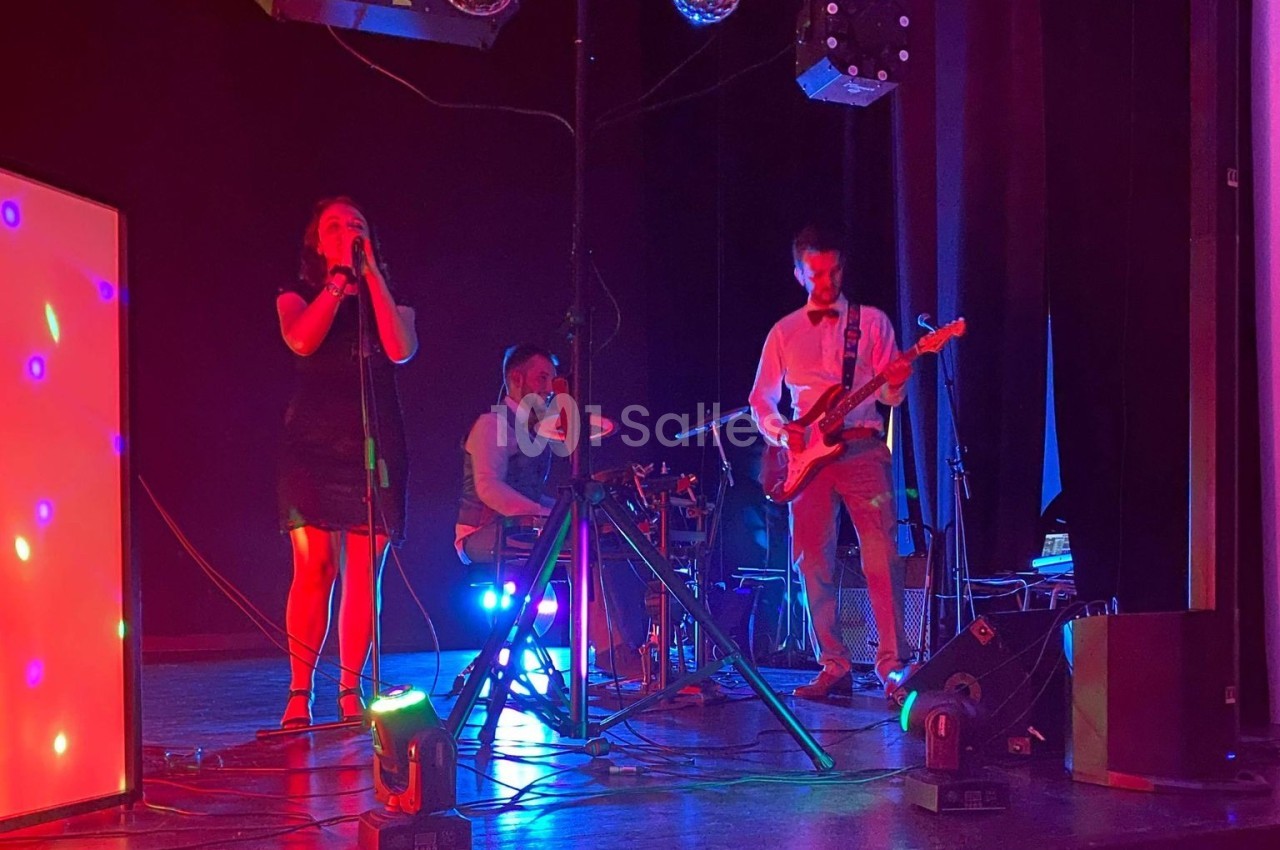 Un groupe de musiciens joue sur scène avec éclairages colorés, comprenant une chanteuse, un batteur et un guitariste.