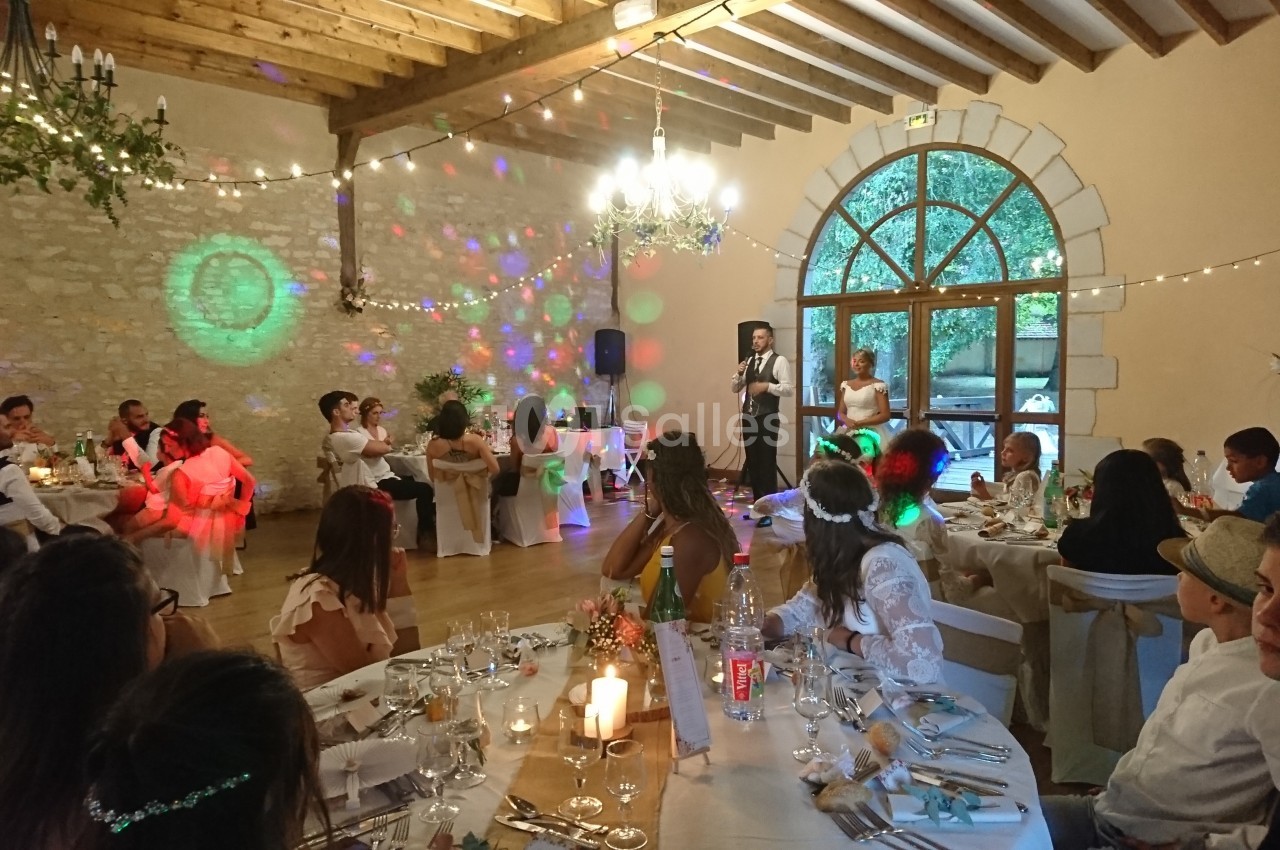 Salle de réception décorée pour un mariage, avec invités assis à table et un couple faisant un discours.