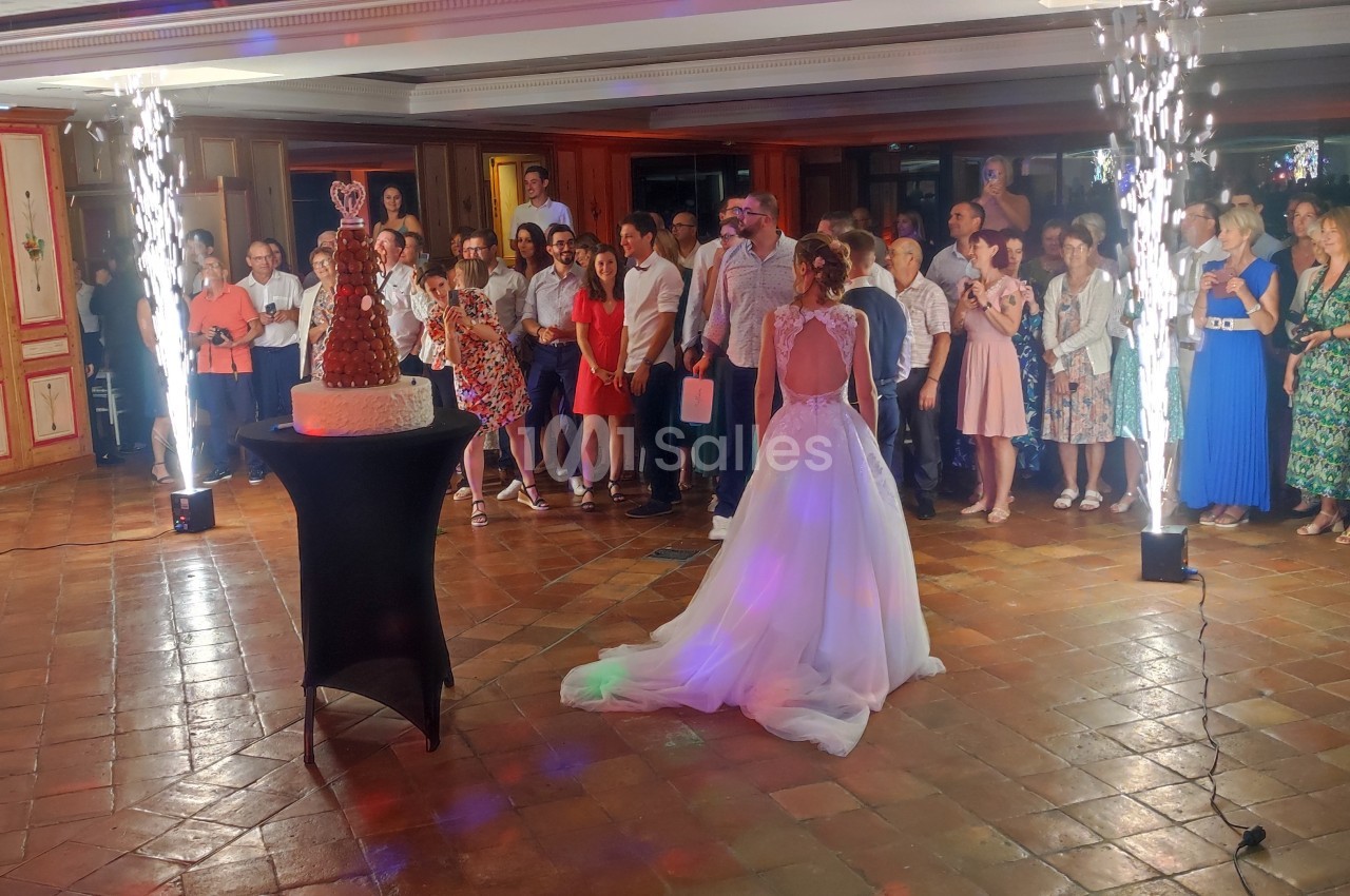 Une mariée en robe blanche regarde un gâteau de mariage entouré d'invités dans une salle décorée avec des étincelles.