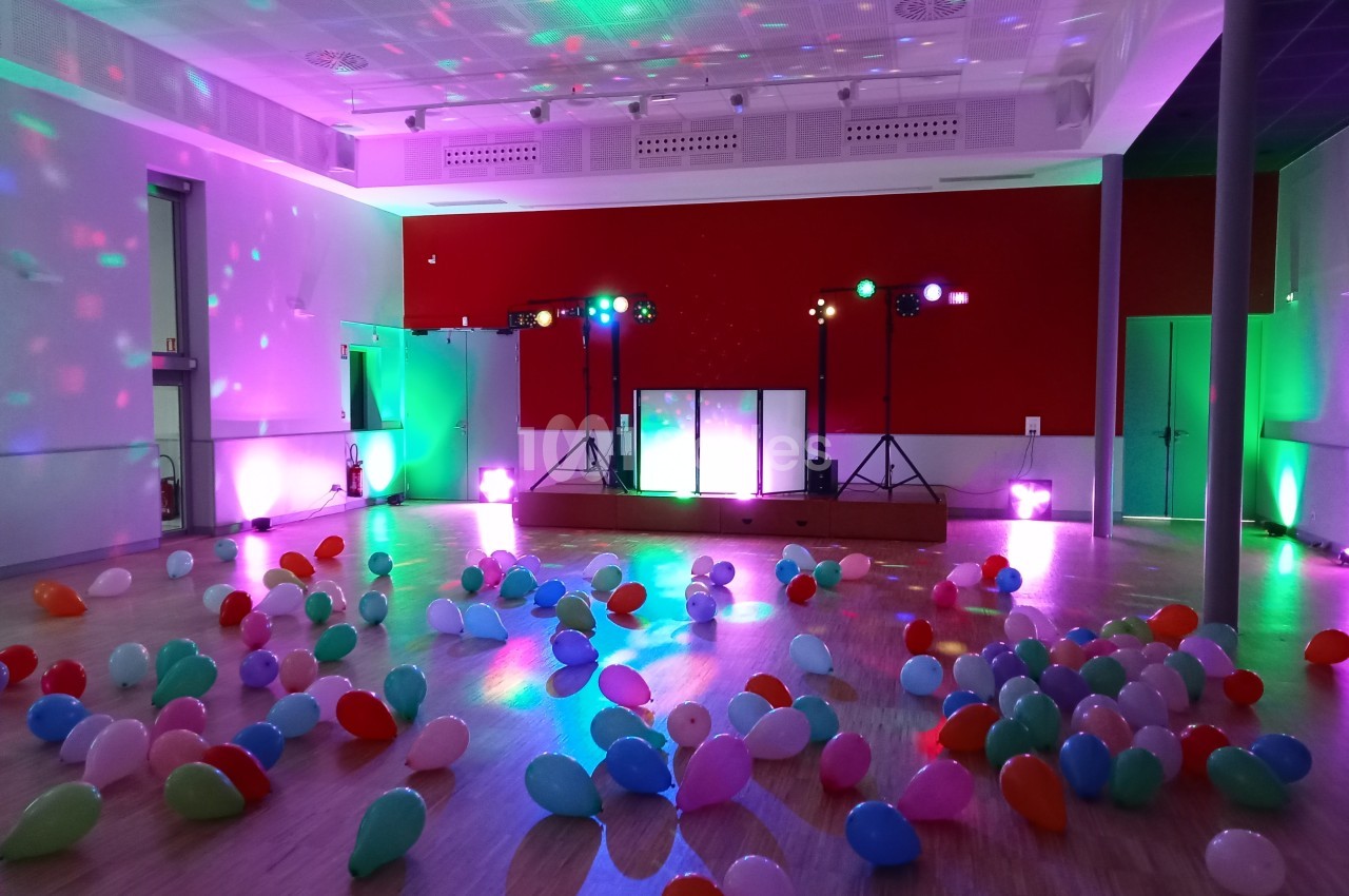 Salle de fête décorée avec des ballons colorés au sol et des lumières d'ambiance sur une scène équipée.